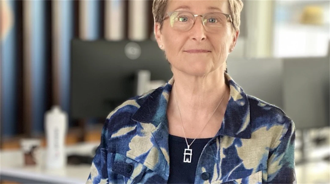 Kvinna med kort brunt hår och glasögon och blå klöder, sitter på en stol i ett kontorslandskp