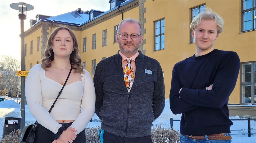 Tre personer står framför en gul byggnad med vita fönster på en snöig dag