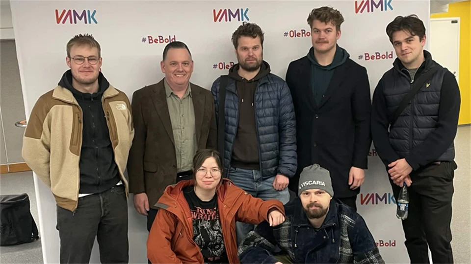 Sju personer poserar för en gruppbild framför en vit bakgrund med VAMK‑logotyp. Fem står upp och två sitter på huk framför dem. Alla bär vardagliga ytterkläder i en inomhusmiljö.