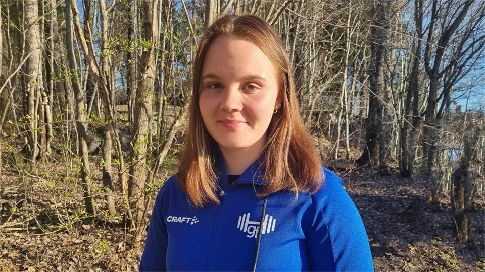 Porträttbild utomhus. En ung tjej med blont hår och i blå tröja står framför en skogsdunge