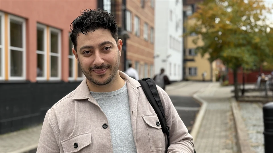 En man tittar in i kameran och ler. I bakgrunden syns några husfasader och träd.