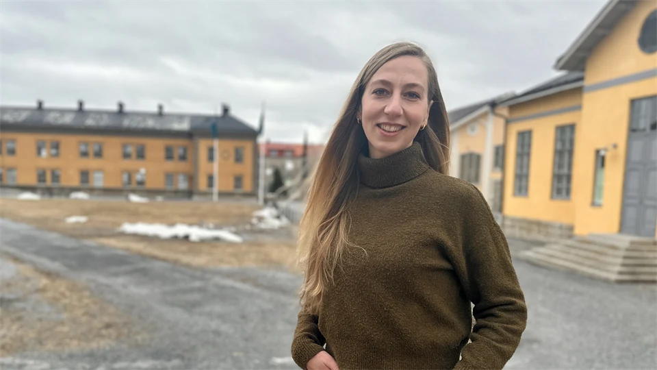 Woman in brown polo shirt and black trousers standing outdoors in front of large yellow buildings