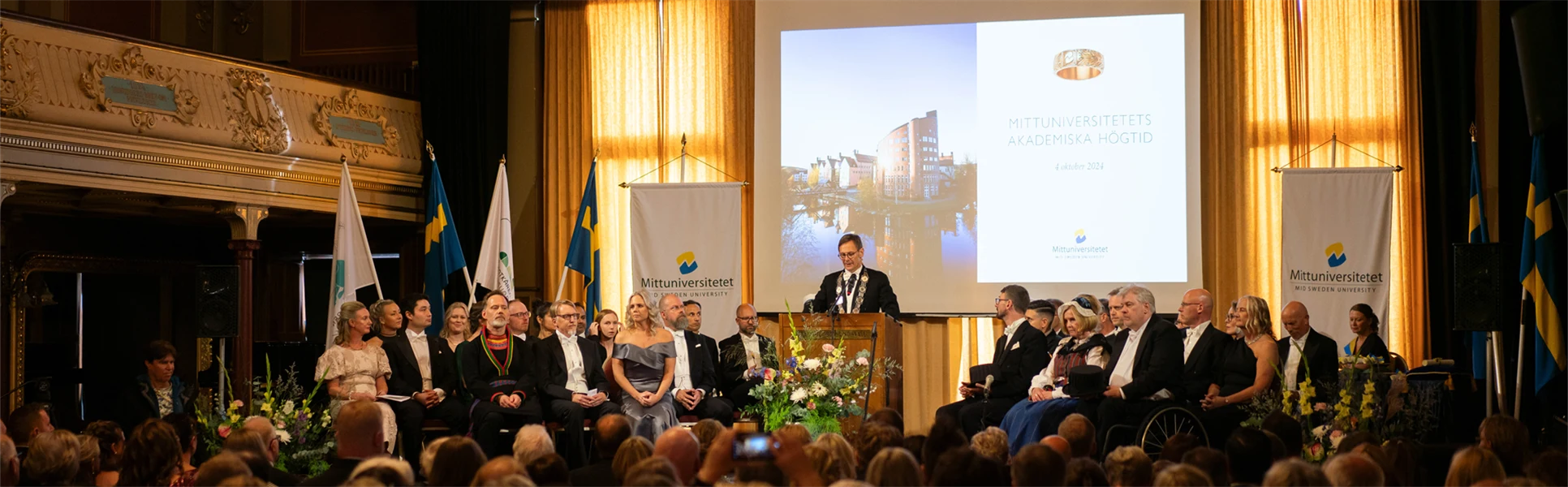 En finklädd man står i en talarstol och talar framför en publik.
