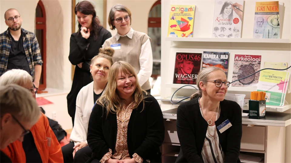 Åtta personer i biblioteksmiljö, några sitter och några står upp, och bakom dem finns vägghyllor med böcker. Vissa av personerna har namnskyltar på sig.   