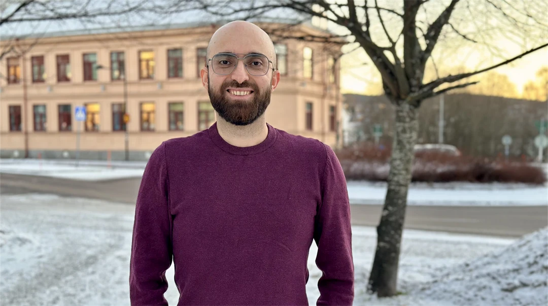 Smiling man with a shaved head, brown beard, and glasses. He is wearing a burgundy sweater and standing outdoors. The season is winter.