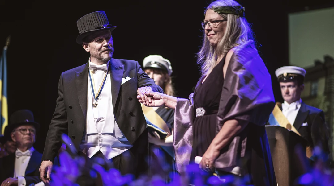 Två finklädda personer - en med doktorshatt, en med lagerkrans håller hand.