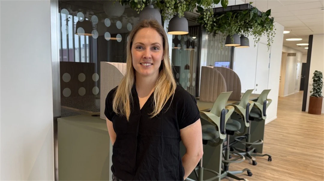 En kvinna tittar in i kameran och ler. I bakgrunden syns ett kontorslandskap.