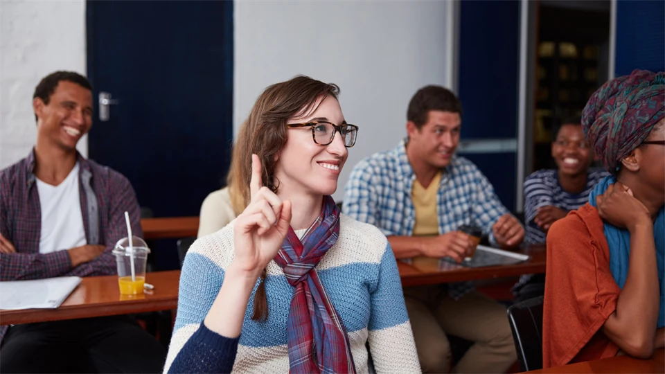 Female student putting her hand up in the classroom to ask a question. There are more students in the classroom and everybody sits at their desks.