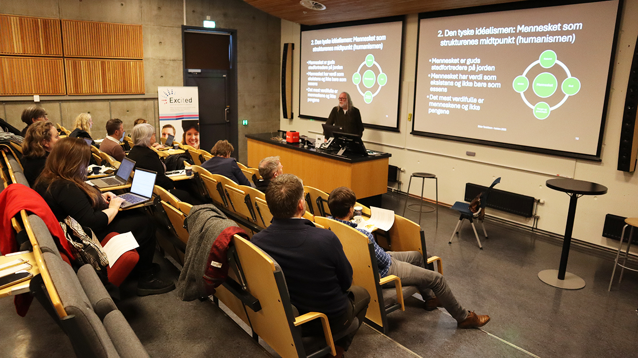 En föreläsningssal där en man står längst fram i salen framför två skärmar med en presentation på och i salen sitter ett tiotal personer i gradäng. 