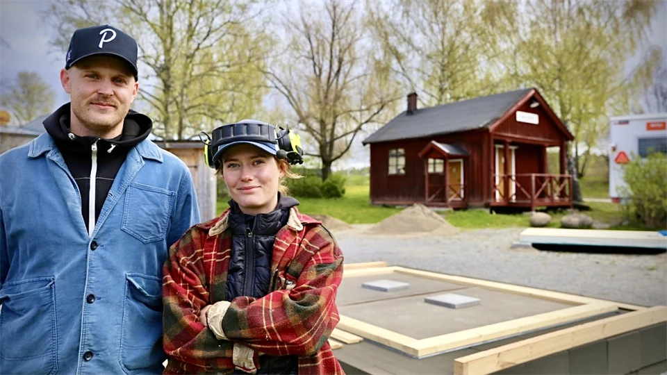 Två personer, en man och en kvinna, står utomhus med två små husgrunder bakom sig. 