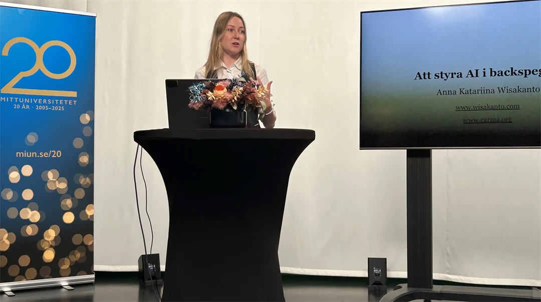 En blond kvinna står vid ett podium. I bakgrunden syns en stor tv-skärm med en presentation på. 