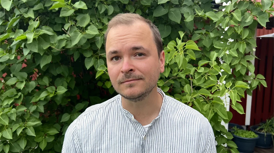 En man står framför en grön buske och tittar in i kameran.