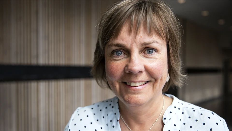 Kvinna med kortklippt frisyr och prickig blus står framför en vägg med träpanel och ler.  