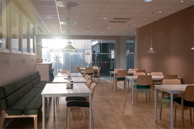 Upplyst sal med ljust interiör fylld med stolar och vita bord, två bänkar med grön klädsel mot väggen till vänster, stor vägg av glasparti i slutet av salen.