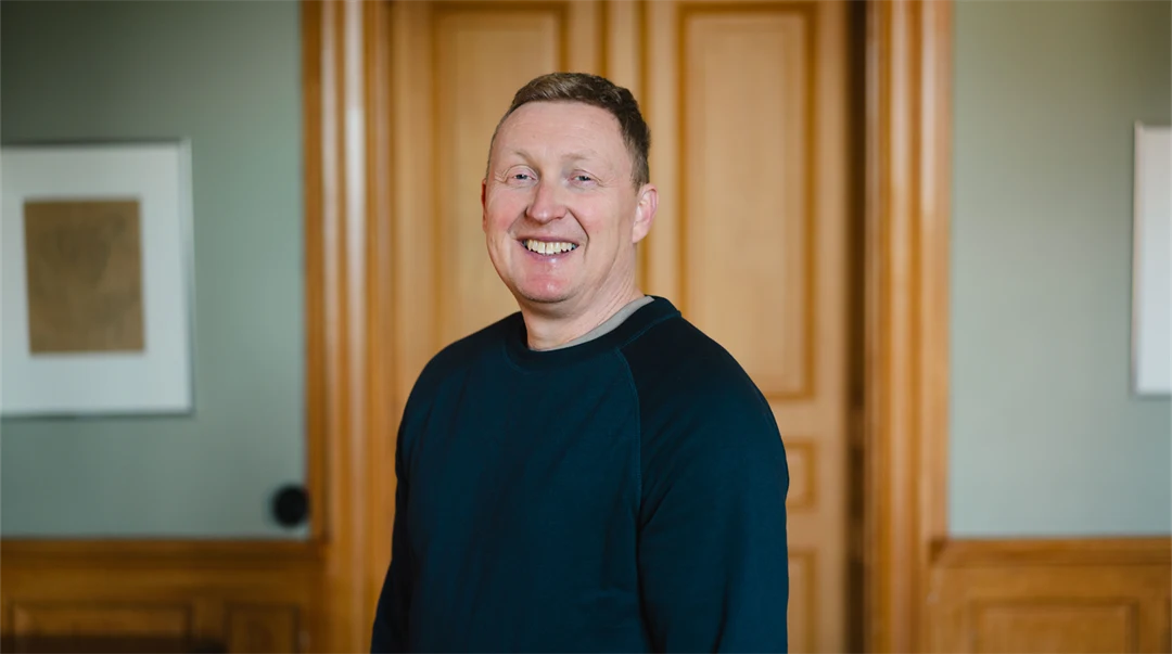 Man i blå collegetröja står framför en brun trädörr och ler mot kameran. 