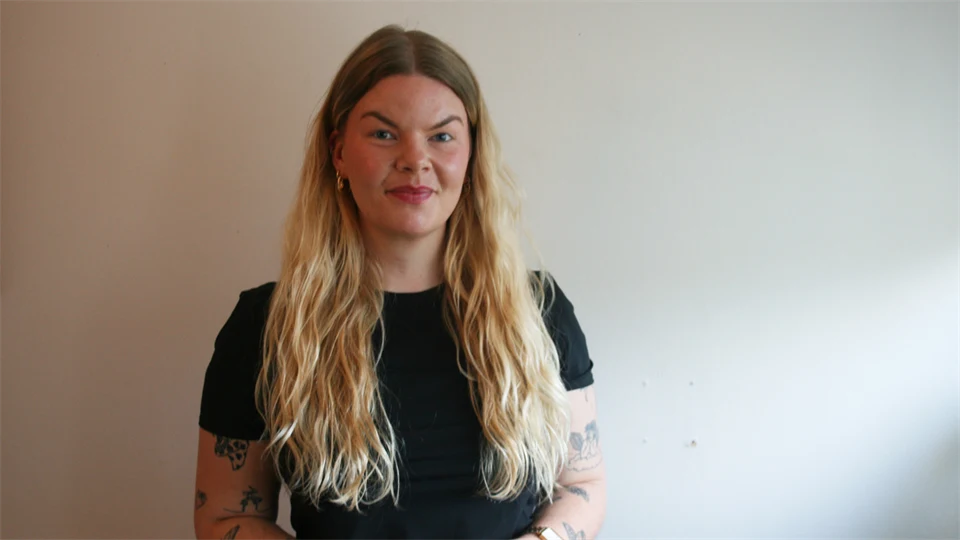 Kvinna i svart kortärmad tröja och långt blont hår står framför en beige vägg. 