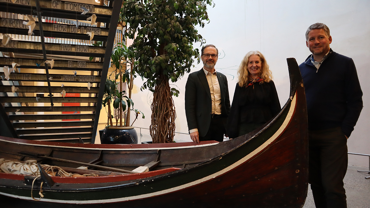 Tre personer, två män och en kvinna står bakom en kanotliknande båt i inomhusmiljö. 