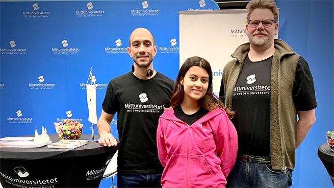 Tre studenter står bredvid varandra på rad till höger om ett bord med broschyrer framför en utställning med blå skyltning.
