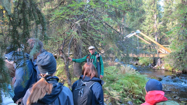 En grupp människor i friluftskläder står i skog och lyssnar på en man som pratar.