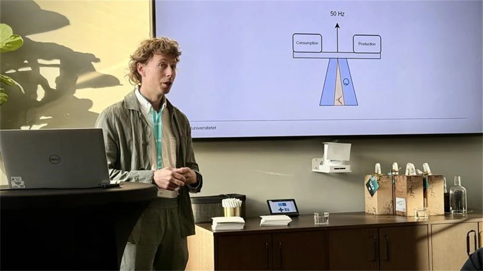 En man med ljusbrun kostym och vit skjorta pratar framför en skärm