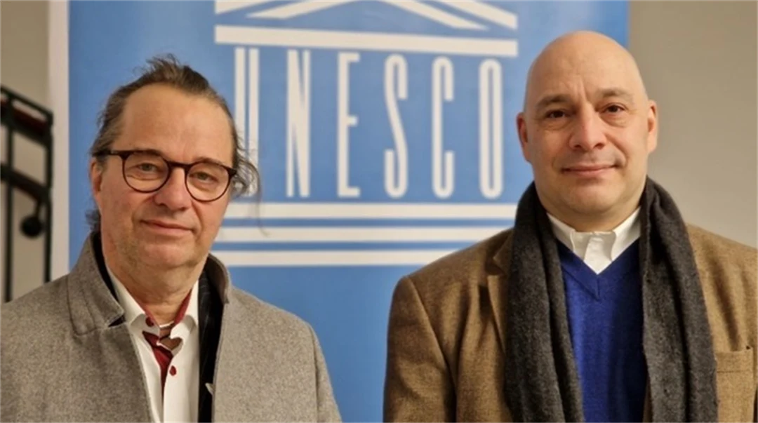 Två män klädda i rockar står framför en blå skylt med UNESCO-logotyp 