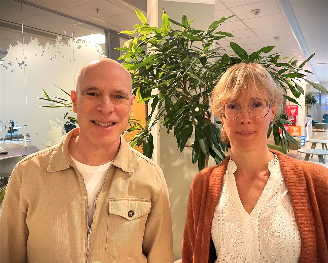 En bild på en man och kvinna från Folkhälsomyndigheten. Mannen står till vänster och kvinnan till höger. I bakgrunden finns et kontorslandskap och gröna växter