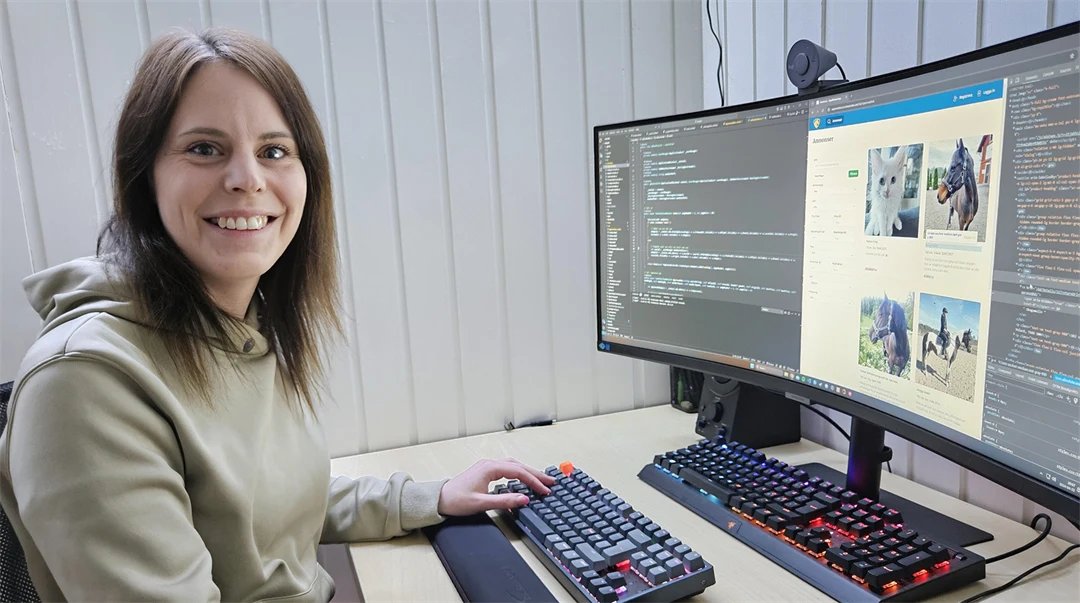 En kvinna i grå tröja sitter vid en arbetsplats med två bildskärmar framför sig. Hon ler och tittar in i kameran.