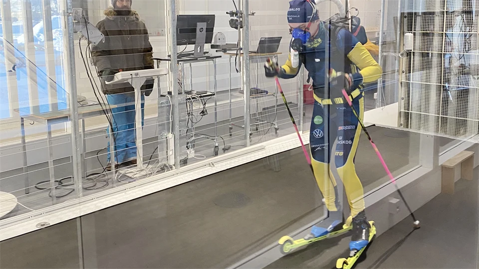 Man i blågul dräkt på rullskidor i vintunneln. En annan man står utanför vindtunneln.