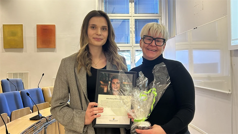 Två kvinnor står tillsammans i en föreläsningssal och håller en blombukett och en plakett i handen.