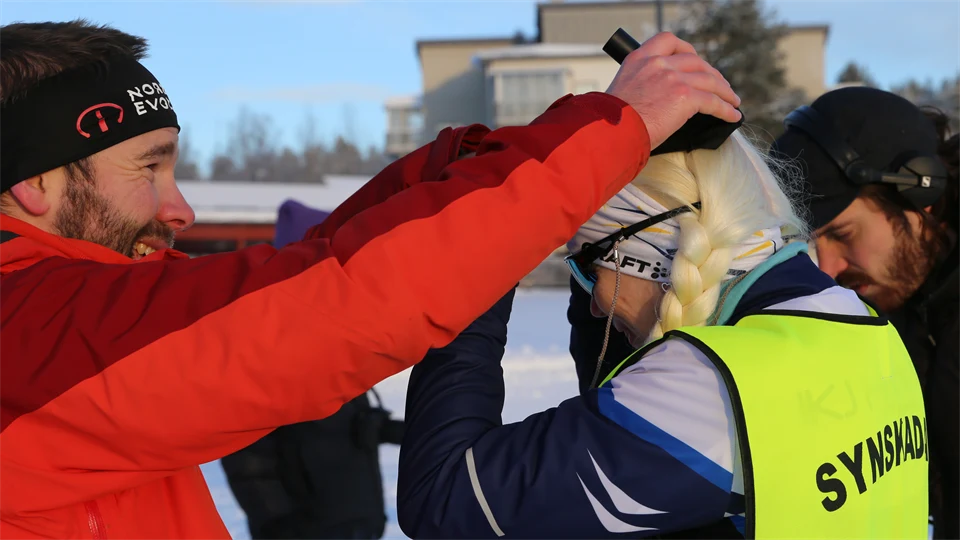 Man i röd jacka hjälper synskadad, kvinnlig skidåkare att bära skallbenshörlurar 