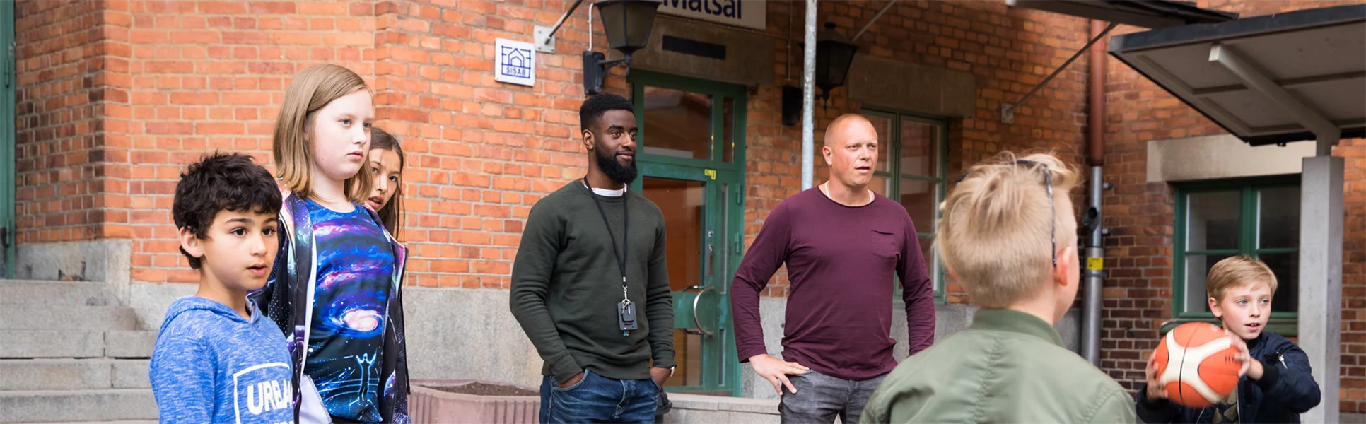 Barn leker med boll på skolgård, två vuxna närvarande.