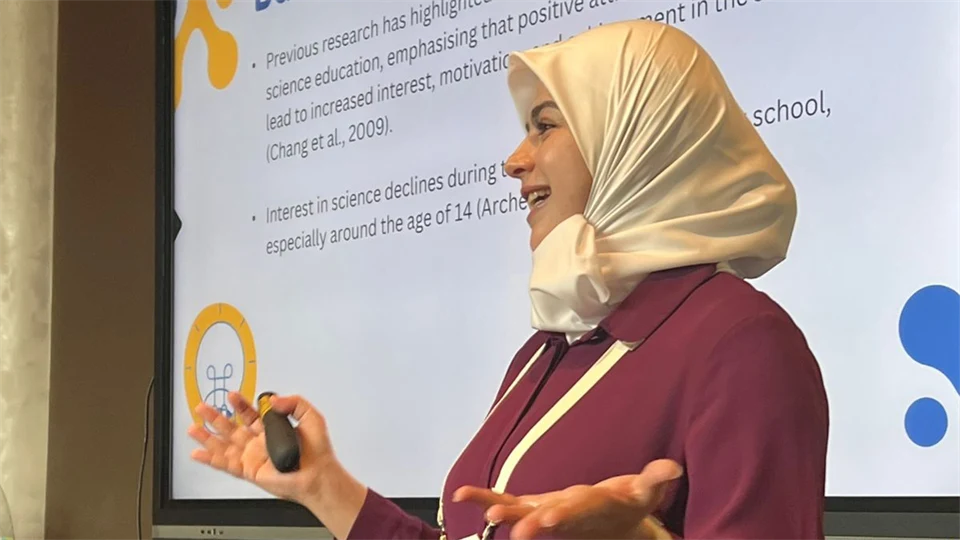 A woman in hijab with a large monitor in the background.