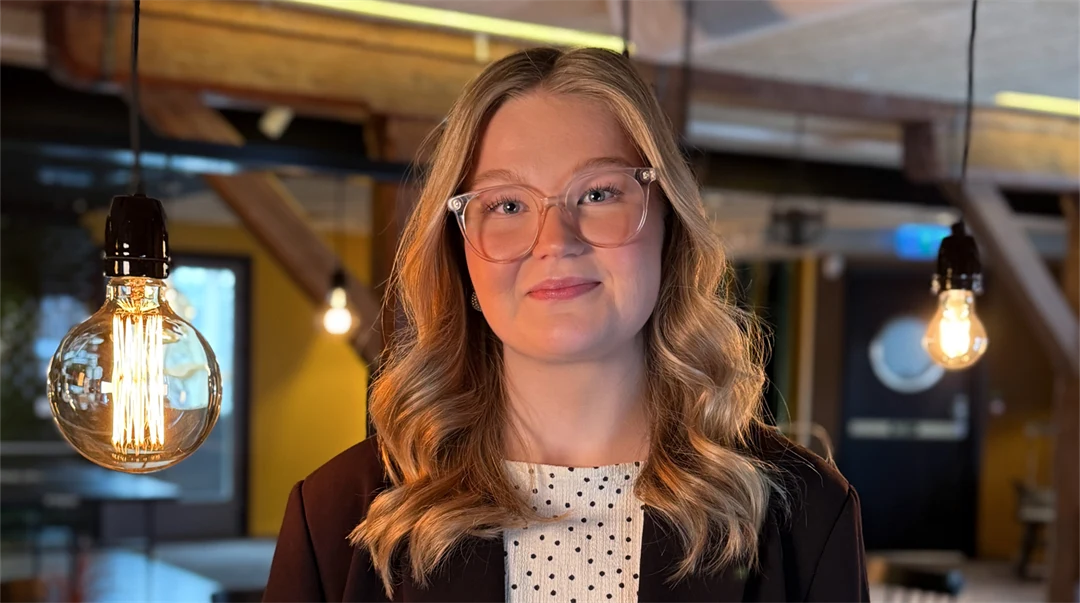 En kvinna ser in i kameran och ler. Lampor hänger i bakgrunden.