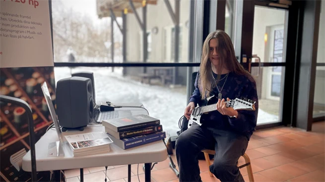 En person med långt hår ler mot kameran och sitter på en stol med en gitarr.