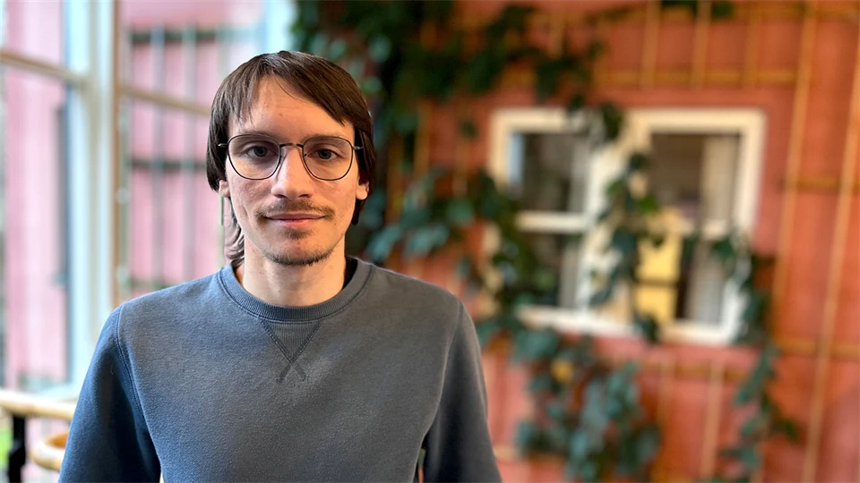 A man with brown hair, glasses and a grey jumper stands in front of a reddish-brown wall covered in climbing green plants. 