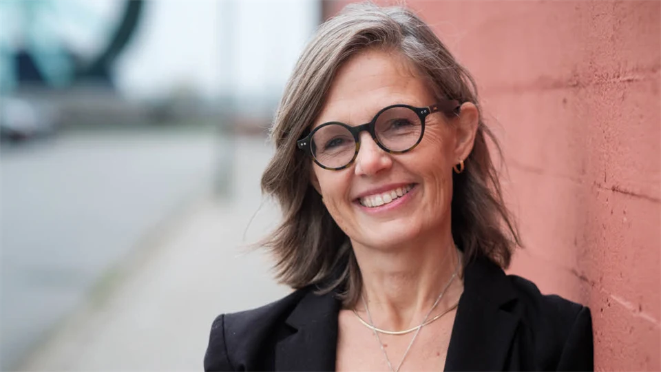 En svartklädd Susanna Hedenborg med glasögon står mot en husfasad och ser glad ut