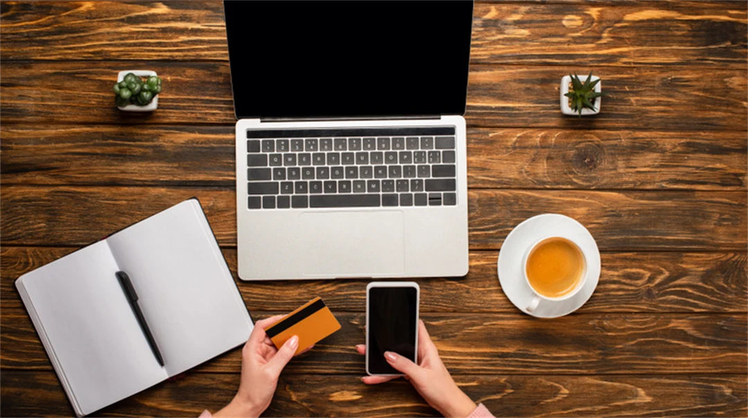 Bord med en laptop, kaffekopp ett uppslaget block och en kvinna som håller i en telefon och ett betalkort.