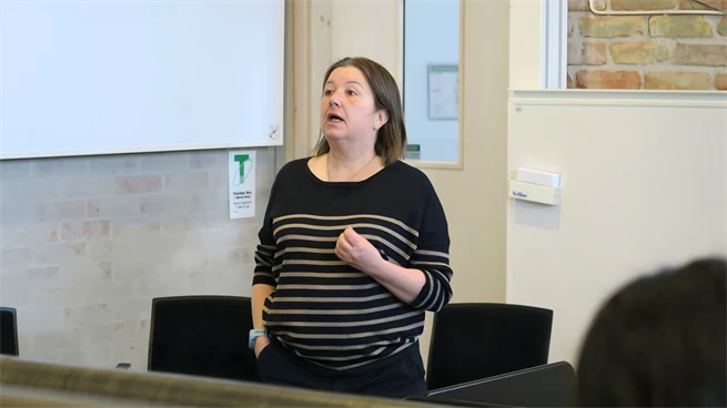 Kvinna står längst fram i en aula och pratar med åhörarna. Hon bär en mörk tröja med vita ränder.