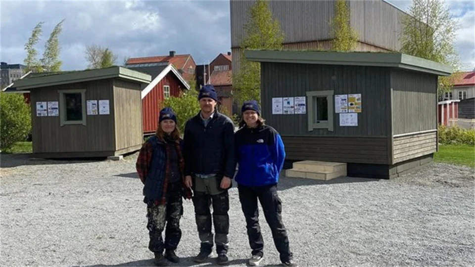 Three persons are standing outside in front of two small buildings.