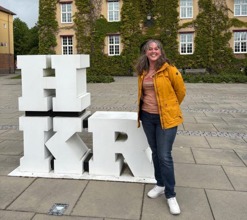 A woman in a yellow jacket is standing outside a beige building with green climbing plants. Next to her is a sculpture with the letters ‘HKR’.