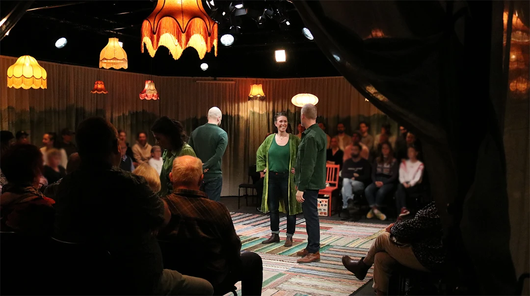 En teaterscen med tre personer i gröna tröjor på och publik runtomkring. 