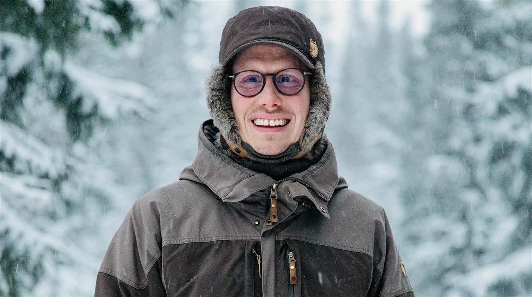 Happy young man in thick winter clothes standing outdoors in a winter landscape with snow-covered fir trees in the background.