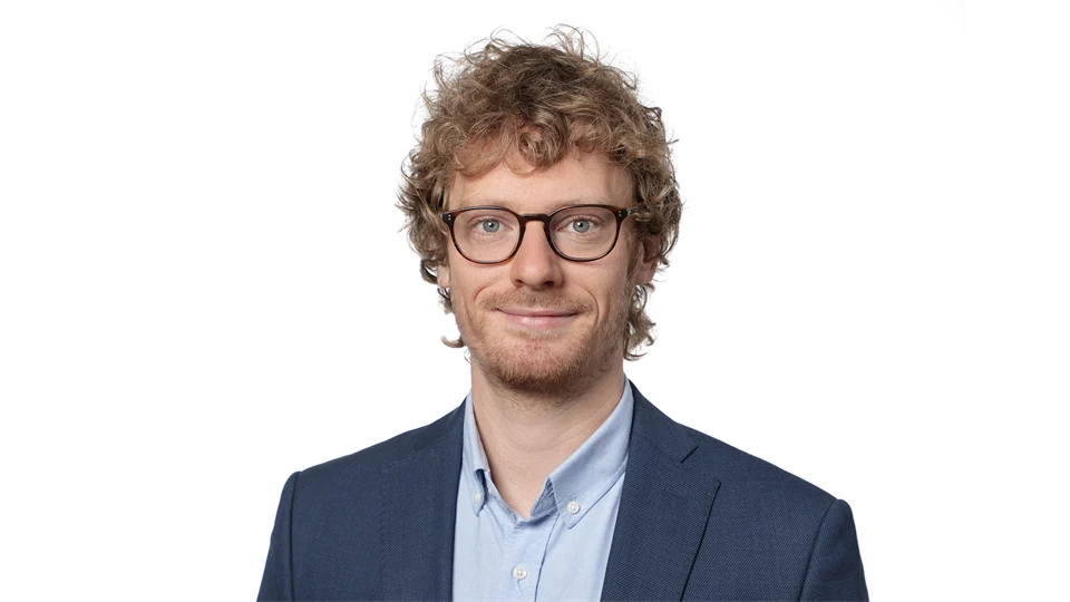 A man with curly hair, glasses, wearing a light blue shirt and dark blue jacket.