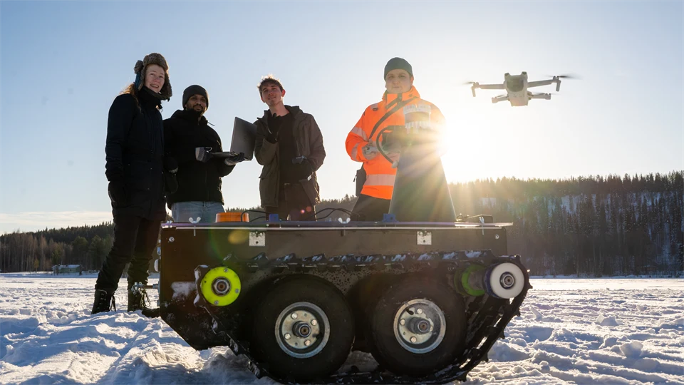  Four people are standing in a snowy landscape. A drone flies behind them and a small all-terrain vehicle stands in front of them.