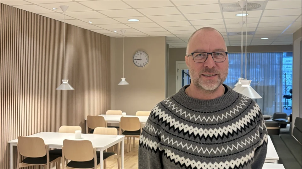 Man in a gray knitted sweater with glasses smiles at the camera, a hall with bright interior, chairs, and white tables in the background.