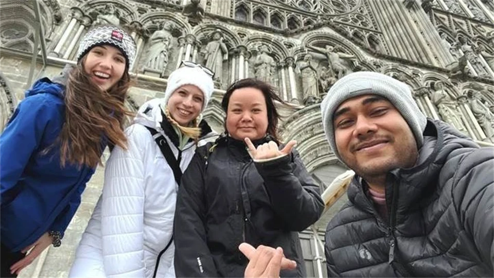Four happy, young people are standing in front of a church wearing outerwear.