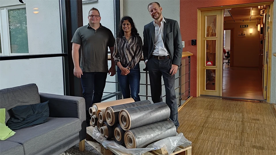  Three people stand together behind a stack of silver-grey large rolls of paper.