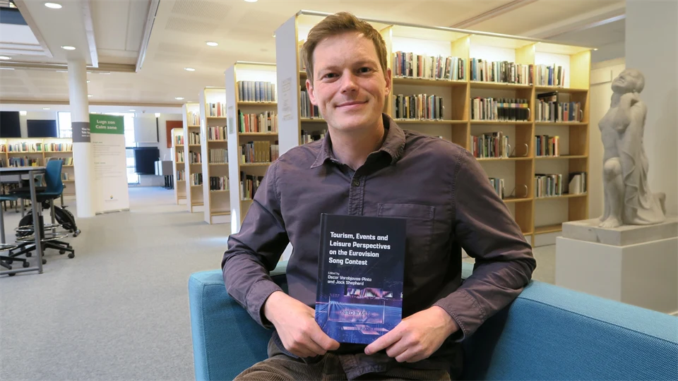 Ung man sittandes i ett bibliotek hållandes en bok. Han har en kort frisyr och bär grå skjorta. 