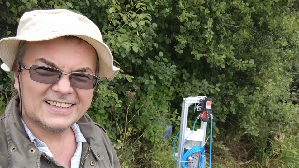 A man in a white hat stands in front of a grove of trees, in the background is measuring equipment.