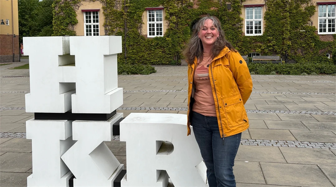 En kvinna i gul jacka står utanför en beige byggnad med gröna klätterväxter på. Bredvid henne står en skulptur med bokstäverna "HKR".